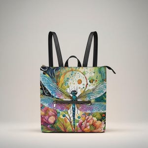 May include: Square backpack with a colourful dragonfly design. The bag features a large dragonfly with blue, purple, and green wings, set against a watercolour background with flowers. It has black straps and a zip closure.