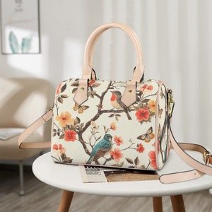 Op de afbeelding: Cr&egrave;mekleurige handtas met een bloemen- en vogelprint. De tas heeft een handvat aan de bovenkant en een afneembare schouderriem. Het ontwerp bevat rode, oranje en gele bloemen, vlinders en vogels op takken. De handvatten en riem zijn lichtroze.