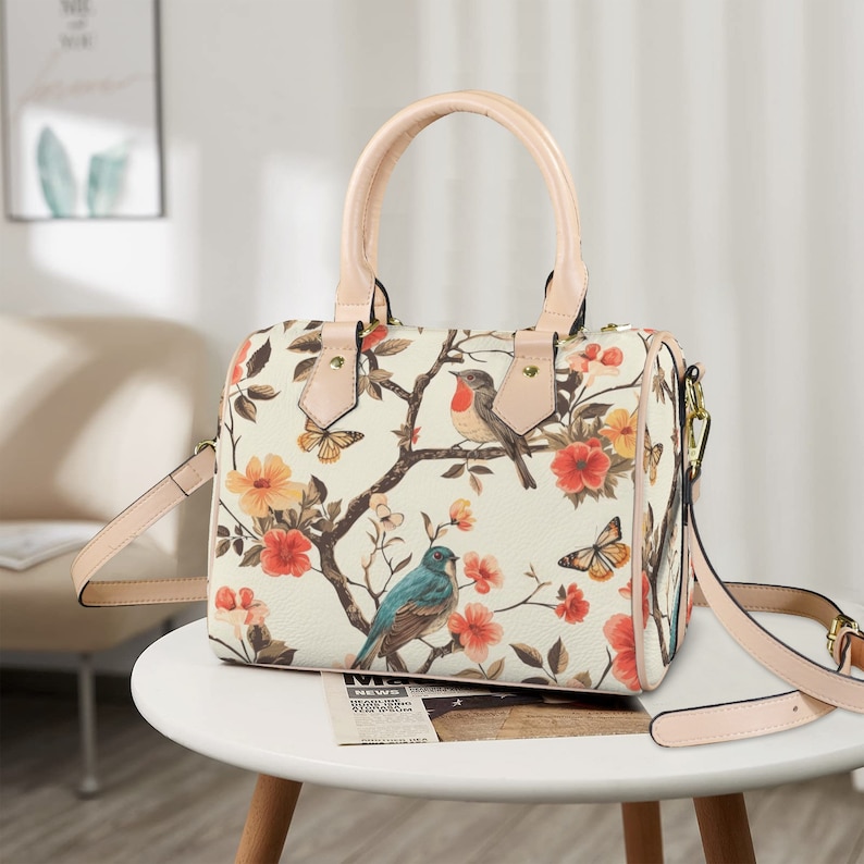 Op de afbeelding: Cr&egrave;mekleurige handtas met een bloemen- en vogelprint. De tas heeft een handvat aan de bovenkant en een afneembare schouderriem. Het ontwerp bevat rode, oranje en gele bloemen, vlinders en vogels op takken. De handvatten en riem zijn lichtroze.