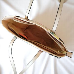 Puede incluir: Una bolsa de mano blanca con estampado floral y una cremallera dorada. La bolsa tiene dos asas y un bolsillo con cremallera en el interior.