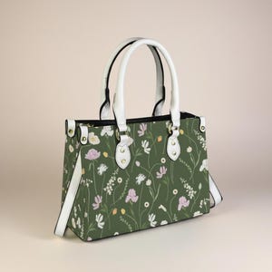 Op de afbeelding: Een groene handtas met een bloemenpatroon van witte, roze en gele bloemen. De tas heeft witte handvatten en een witte schouderriem. Goudkleurige hardware accentueert het ontwerp van de tas.