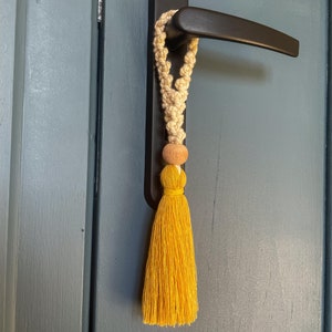 Op de afbeelding: Een decoratieve deurklink kwast. De kwast is gemaakt van een geweven beige koord, een houten kraal en een felgele kwast. De handgreep is zwart en gemonteerd op een blauwe deur.