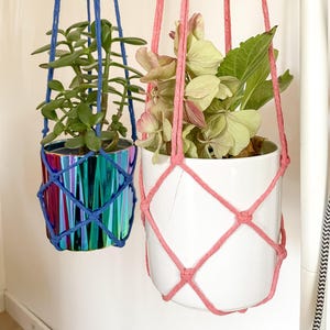 Pode incluir: Duas floreiras suspensas. Uma é de cor arco-íris metálica, suspensa por macramé azul. A outra é um vaso de cerâmica branco com macramé rosa. Ambas são suspensas por cima.