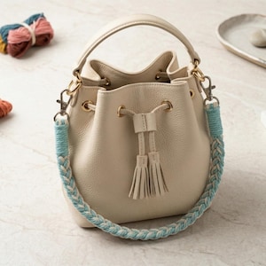 May include: Cream-colored bucket bag with a braided blue and white strap. The bag has a top handle and drawstring closure with tassels. Gold-tone hardware accents the bag's design. The bag is made of a textured material.