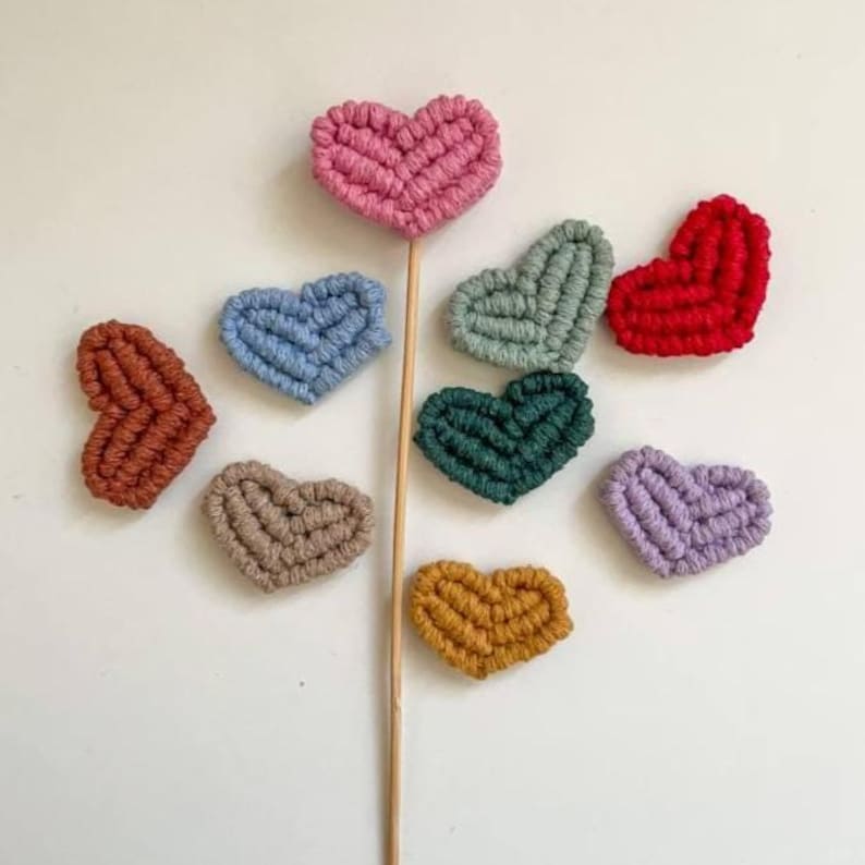 May include: A selection of handmade, colourful, crocheted hearts in shades of pink, blue, green, red, brown, tan, yellow, and purple. One heart is attached to a wooden stick. The hearts are arranged on a white surface.
