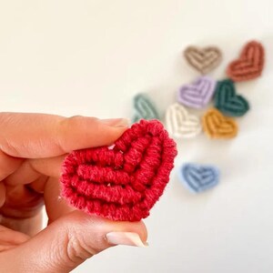 May include: A close-up of a red, heart-shaped, handmade item. The item is made of a textured, woven material. Several other heart-shaped items in various colours are in the background, including beige, green, purple, and blue.