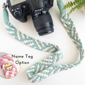 Puede incluir: Una correa trenzada para cámara en azul claro y crema, sujeta a una cámara Nikon negra. El diseño de la correa es visible. Un primer plano muestra una opción de etiqueta con el nombre "Jeannette" en una etiqueta de madera.
