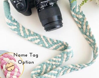 May include: A braided camera strap in a pattern of light blue and cream. The strap is attached to a black Nikon camera. A name tag option is shown in a close-up, with the name "Jeannette" on a wooden tag.