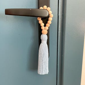 Könnte beinhalten: Ein dekorativer Türgriff-Akzent mit einer Kette aus hellbraunen Holzperlen und einer hellblauen Quaste. Der Akzent hängt an einem schwarzen Türgriff an einer blauen Tür.