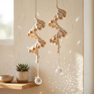 Op de afbeelding: Twee macramé hangdecoraties met houten kralen en kristallen hangers. Het gebroken witte macramé is gerangschikt in een zigzagpatroon, met houten kralen gegroepeerd langs het ontwerp. Heldere kristallen hangers hangen van onderen.