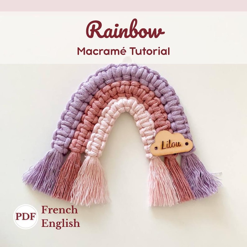 K&ouml;nnte beinhalten: Ein handgefertigter Makramee-Regenbogen in Lila-, Rosa- und Pfirsicht&ouml;nen, mit einem Holz-Wolkenanh&auml;nger mit der Aufschrift "Lilou". Der Regenbogen hat eine strukturierte Optik. Der Text "Rainbow Macram&eacute; Tutorial" ist ebenfalls vorhanden.