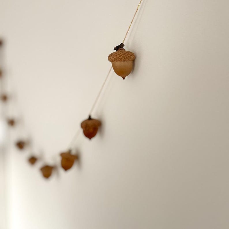 May include: A garland of small, brown acorn ornaments hangs against a white wall. The acorns are strung together with a thin, gold-colored string. The acorns have a natural, rustic appearance, with detailed caps and smooth bodies.