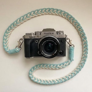 May include: A silver and black Fujifilm X-T4 camera with a lens is shown with a braided camera strap. The strap is a combination of white and light blue. The camera strap is attached to the camera with silver metal clasps.