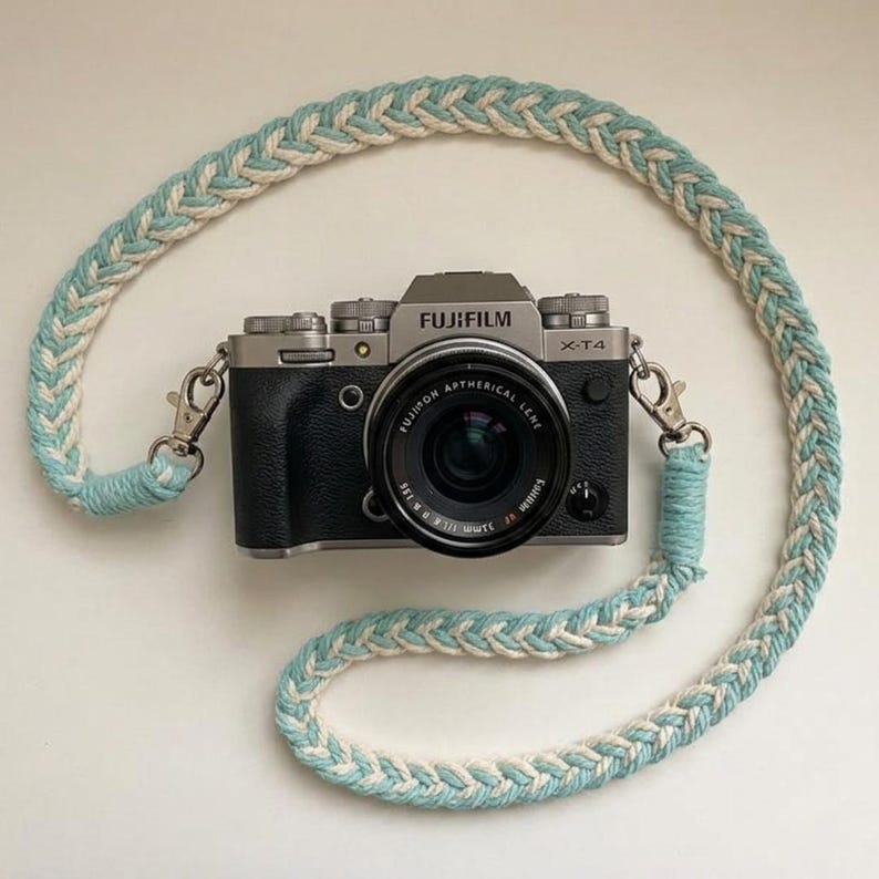 May include: A silver and black Fujifilm X-T4 camera with a lens is shown with a braided camera strap. The strap is a combination of white and light blue. The camera strap is attached to the camera with silver metal clasps.