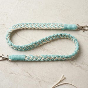 May include: Braided lanyard in a light blue and cream color scheme. The lanyard is made of thick, woven rope and features silver-colored metal swivel clasps on each end. The ends of the lanyard are wrapped in solid light blue thread.