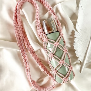 May include: A light green water bottle with a silver lid is held in a pink macrame bottle holder. The holder has a woven net design and a long strap. The background is a white fabric with a decorative white feather.
