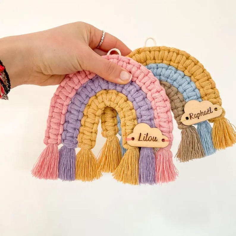 K&ouml;nnte beinhalten: Zwei handgefertigte Makramee-Regenbogen-Dekorationen in Pastellfarben. Einer ist rosa, lila und gelb, mit einem Holz-Wolkenanh&auml;nger mit dem Namen "Lilou". Der andere ist gelb, blau und braun, mit dem Namen "Rapha&euml;l". Jeder Regenbogen hat passende Quasten.