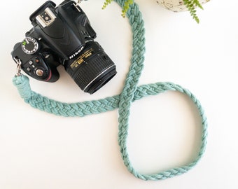 May include: A black Nikon DSLR camera with a mint green braided camera strap. The strap is made of a thick, woven material. The camera lens is black with gold lettering. The camera is on a white surface.