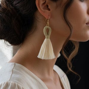 Puede incluir: Pendientes de borla color crema con detalles metálicos dorados. Los pendientes presentan un diseño en bucle en la parte superior, que conduce a una borla larga y fluida. Los pendientes están diseñados para usarse con un gancho.