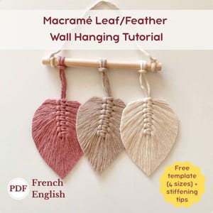 May include: A macramé wall hanging tutorial featuring three leaf-shaped decorations in shades of pink, beige, and cream. The leaves are suspended from a wooden dowel. The image includes text that reads "Macramé Leaf/Feather Wall Hanging Tutorial".
