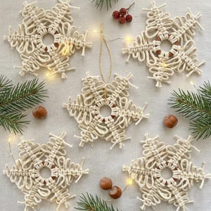 Puede incluir: Cinco adornos de macram&eacute; en color blanco roto con forma de copo de nieve, con intrincados patrones de cord&oacute;n anudado alrededor de anillos de madera. Cada decoraci&oacute;n en forma de estrella incluye un lazo para colgar y ramas de pino y avellanas.
