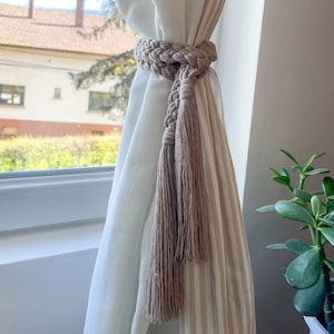May include: A beige macrame curtain tieback with a braided design and long tassels. The tieback is wrapped around a white curtain with vertical stripes, creating a decorative element for window treatments. The natural tones complement the neutral color palette.