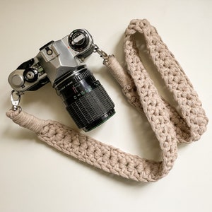 Può includere: Una fotocamera vintage argento e nera con un obiettivo nero e una tracolla beige all'uncinetto. La tracolla è realizzata in filato spesso e ha chiusure in metallo. La fotocamera è su una superficie bianca.