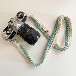Op de afbeelding: Een zilveren filmcamera met een zwarte lens is bevestigd aan een crème- en zeegroene macramé camerariem. De riem heeft een klein houten label met de tekst "Ambre".