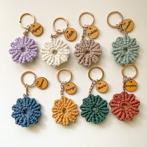 Peut inclure: Plusieurs porte-clés faits à la main en forme de fleurs au crochet, de différentes couleurs : violet, blanc, marron, vert, bleu, jaune et rouge. Chaque porte-clé est orné d'un anneau doré et d'une petite étiquette en bois gravée d'un nom.