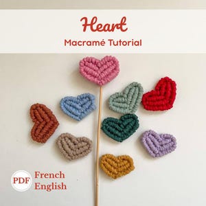 Peut inclure: Plusieurs coeurs en macramé de différentes couleurs, dont rose, bleu, vert et rouge. Un coeur est monté sur une baguette en bois. Le texte "Heart Macramé Tutorial" est également présent.