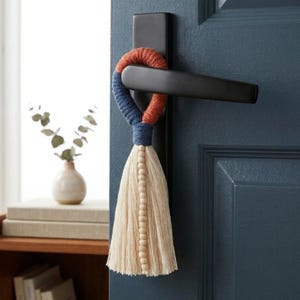 Peut inclure: Un pompon de porte décoratif avec un design tissé en orange, bleu et crème. Le pompon est orné de perles en bois et est suspendu à une poignée de porte noire. La porte est peinte en bleu foncé.