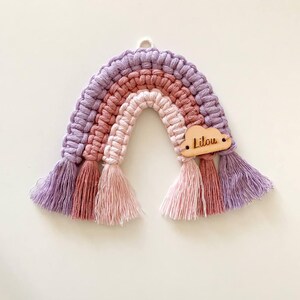 K&ouml;nnte beinhalten: Handgefertigter Makramee-Regenbogen-Wandbehang in Lila-, Rosa- und Pfirsicht&ouml;nen. Der Regenbogen hat eine Holz-Wolke mit dem eingravierten Namen "Lilou". Die Quasten unten verleihen einen dekorativen Touch.