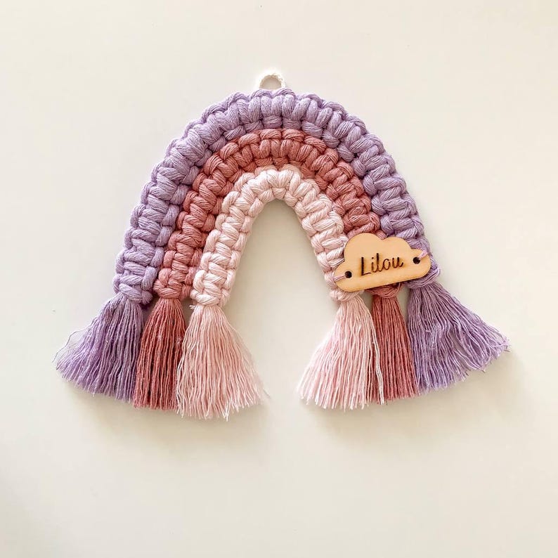 K&ouml;nnte beinhalten: Handgefertigter Makramee-Regenbogen-Wandbehang in Lila-, Rosa- und Pfirsicht&ouml;nen. Der Regenbogen hat eine Holz-Wolke mit dem eingravierten Namen "Lilou". Die Quasten unten verleihen einen dekorativen Touch.
