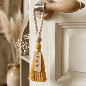 Peut inclure: Un porte-clés en macramé fait à la main avec une perle en bois et un pompon jaune moutarde. Une petite étiquette en bois avec le nom "Émilie" est attachée au pompon. Le porte-clés est suspendu à une poignée de porte en bois.