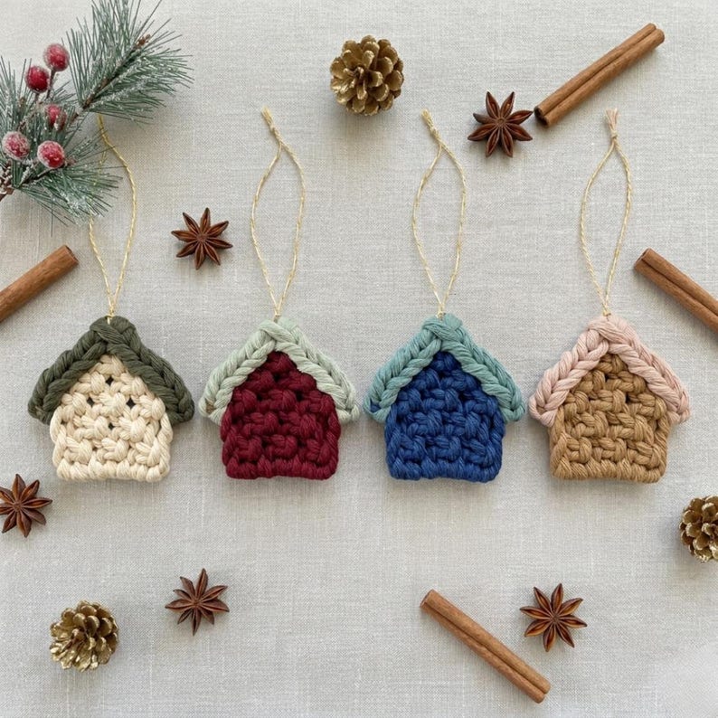 Puede incluir: Cuatro adornos de macram&eacute; en forma de casa con lazos para colgar, con cuerpos tejidos en color crema, burdeos, azul y tostado con techos en contraste. El conjunto est&aacute; dispuesto con agujas de pino, ramas de canela y an&iacute;s estrellado sobre tela.