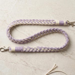 May include: Braided lanyard in lavender and cream. The lanyard is made of thick, woven rope with a repeating pattern. Silver-colored metal clasps are attached at each end. The lanyard is designed to hold keys or an ID badge.