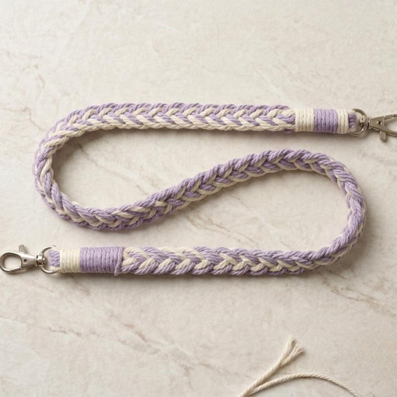 May include: Braided lanyard in lavender and cream. The lanyard is made of thick, woven rope with a repeating pattern. Silver-colored metal clasps are attached at each end. The lanyard is designed to hold keys or an ID badge.