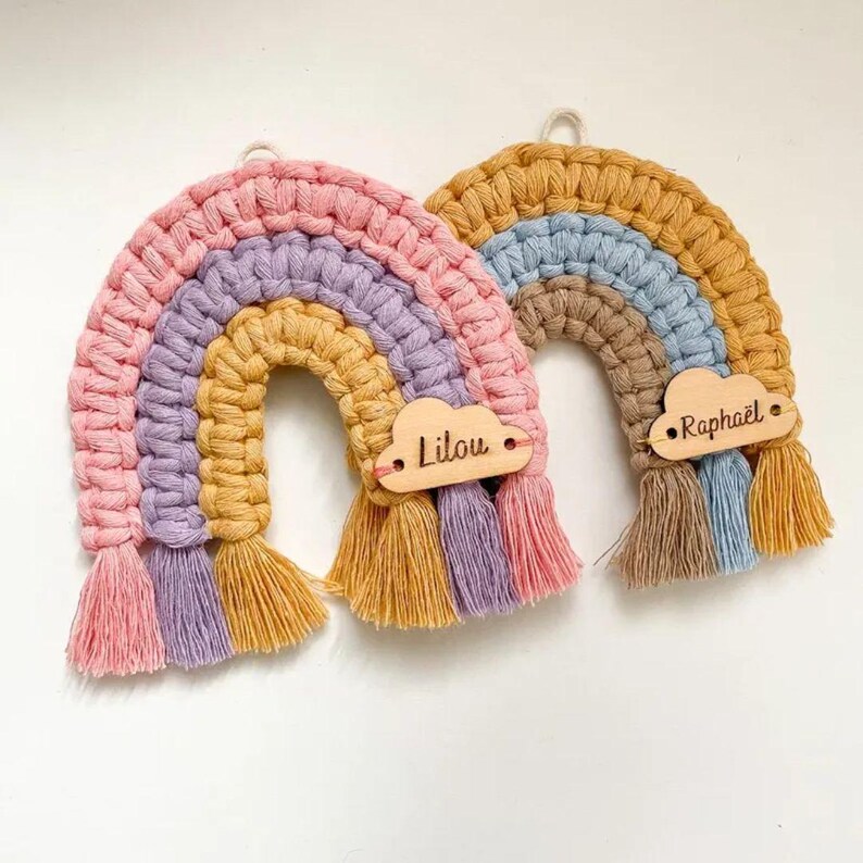 K&ouml;nnte beinhalten: Zwei Makramee-Regenbogen-Wandbeh&auml;nge. Ein Regenbogen ist rosa, lila, gelb und goldfarben. Der andere ist gold, blau, braun und beige. Jeder hat eine Holzwolke mit einem Namen. Der erste hei&szlig;t "Lilou" und der zweite "Rapha&euml;l".