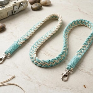 May include: Two braided camera straps. The straps are a combination of white and light blue, with silver metal clasps. The ends of the straps are wrapped in light blue thread. The straps are on a light-colored surface.