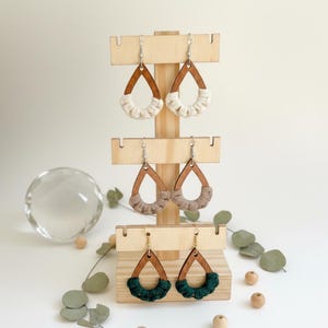 May include: Wooden teardrop earrings with cream, taupe, and green macrame accents are displayed on a wooden earring stand. The earrings have silver-colored hooks. The stand is on a white surface with decorative elements.
