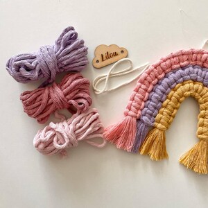 K&ouml;nnte beinhalten: Handgefertigter Makramee-Regenbogen in Rosa, Lila und Gelb mit passenden Baumwollschnurb&uuml;ndeln. Eine kleine Holzwolke mit dem Namen "Lilou" ist ebenfalls vorhanden. Perfekt f&uuml;r die Dekoration im Kinderzimmer.
