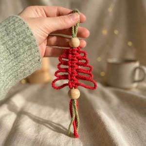 Puede incluir: Un adorno de macram&eacute; con forma de &aacute;rbol de Navidad, elaborado con cuerda roja y detalles en cuerda verde en el lazo superior y el fleco inferior. El dise&ntilde;o presenta dos cuentas de madera claras, redondas y lisas, situadas arriba y abajo del &aacute;rbol.