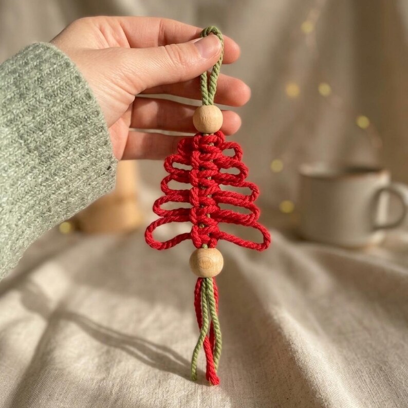 Puede incluir: Un adorno de macram&eacute; con forma de &aacute;rbol de Navidad, elaborado con cuerda roja y detalles en cuerda verde en el lazo superior y el fleco inferior. El dise&ntilde;o presenta dos cuentas de madera claras, redondas y lisas, situadas arriba y abajo del &aacute;rbol.