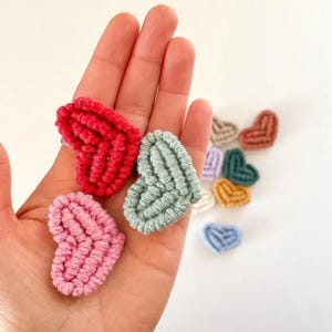 May include: A selection of small, heart-shaped ornaments crafted from textured yarn. The foreground features hearts in red, pink, and green, with others in tan, blue, and yellow visible in the background. These decorative items are handmade.