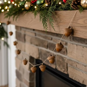 May include: A garland of brown acorns strung on twine hangs in front of a brick fireplace. The acorns are various sizes and have textured caps. The fireplace mantel is decorated with greenery, gold ornaments, and fairy lights, creating a festive holiday display.