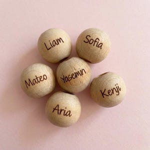Puede incluir: Seis cuentas de madera redondas de color claro, cada una grabada con un nombre único en una fuente marrón oscuro. Los nombres visibles en las cuentas son Liam, Sofia, Mateo, Yasemin, Kenji y Aria. Las cuentas están dispuestas en un grupo.