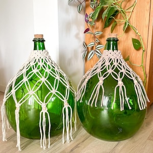 Peut inclure: Deux grandes bouteilles en verre vert avec des bouchons en liège et un filet en macramé crème. Les bouteilles sont rondes avec une base large et un col étroit. Le macramé a un motif en losange et des franges.
