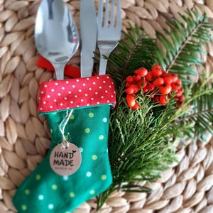 Op de afbeelding: Een feestelijke kerstbestekhouder. De groene kous is versierd met witte stippen en een rode stippenboord. Zilver bestek, waaronder een lepel, mes en vork, is erin geplaatst. Een takje groen en rode bessen maken het feestelijke uiterlijk compleet.