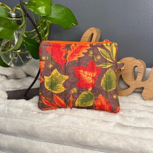 May include: A small, square, quilted coin purse with a brown background and a pattern of autumn leaves in shades of red, orange, and green. The purse has an orange zipper and trim. The overall theme is fall and nature.
