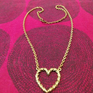May include: A gold necklace featuring a heart-shaped pendant with a twisted rope design. The chain is fine and the necklace is displayed on a pink patterned background.